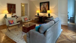 A living room with a grey sofa, two armchairs, a coffee table, and a patterned rug. There are two lamps on the side table and a painting on the wall.