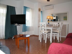 A room with a television, a dining table with chairs, a red sofa, and a kitchen area.