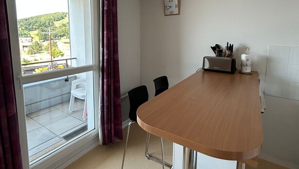 A room with a wooden table, two chairs, a window with curtains, a clock on the wall, and a view of a green landscape outside.