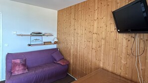 A room with a purple sofa, a mounted TV, wooden panel walls, and a shelf with books and a framed picture.