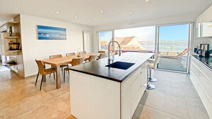 Une cuisine moderne dotée d’un îlot central, d’un coin repas et de grandes fenêtres offrant une vue sur la mer.