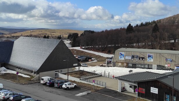 Un bâtiment doté d’un toit en pente, un espace de stationnement avec plusieurs voitures, et une vue sur un paysage enneigé avec des arbres et des montagnes au loin.