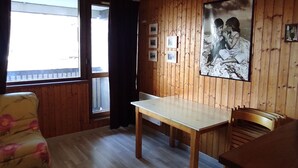 Une pièce aux murs recouverts de lambris en bois, une grande fenêtre avec des rideaux, une table en bois, un fauteuil à motifs floraux et une photographie encadrée accrochée au mur.
