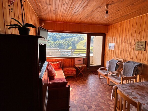 A wooden cabin interior with a sofa, chairs, a table, and a view of a forested area through a large window.