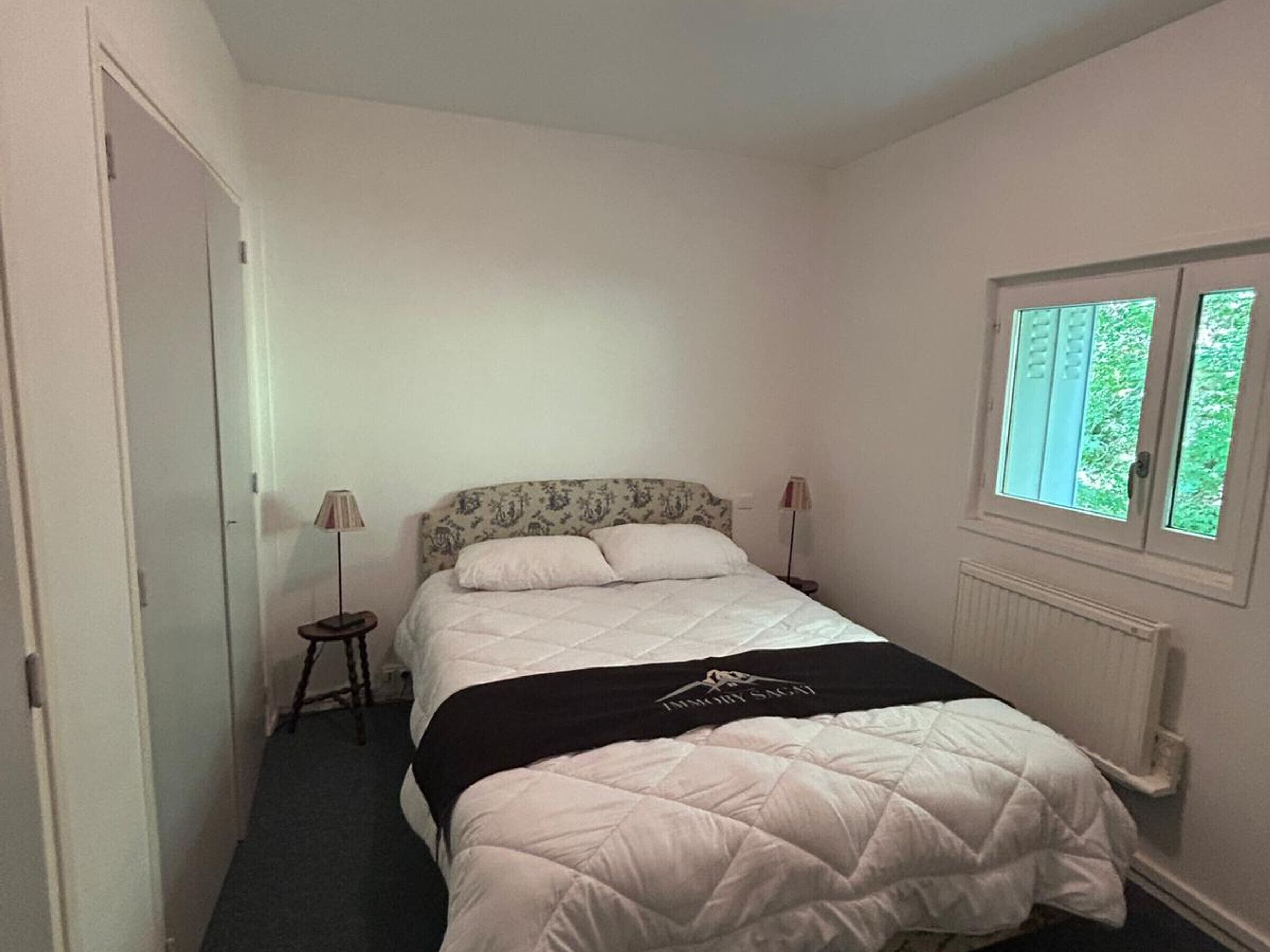 A small, neatly arranged bedroom with a single bed, two bedside lamps, a window with a view of greenery, and a radiator.