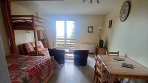 Een kamer met een stapelbed, een bureau met stoelen, een raam met uitzicht en een klok aan de muur.
