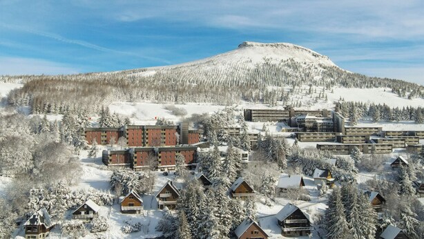 Een met sneeuw bedekt wintersportresort met meerdere gebouwen, waaronder een groot hotel en kleinere huisjes, gelegen voor een besneeuwde berg en dicht bos.
