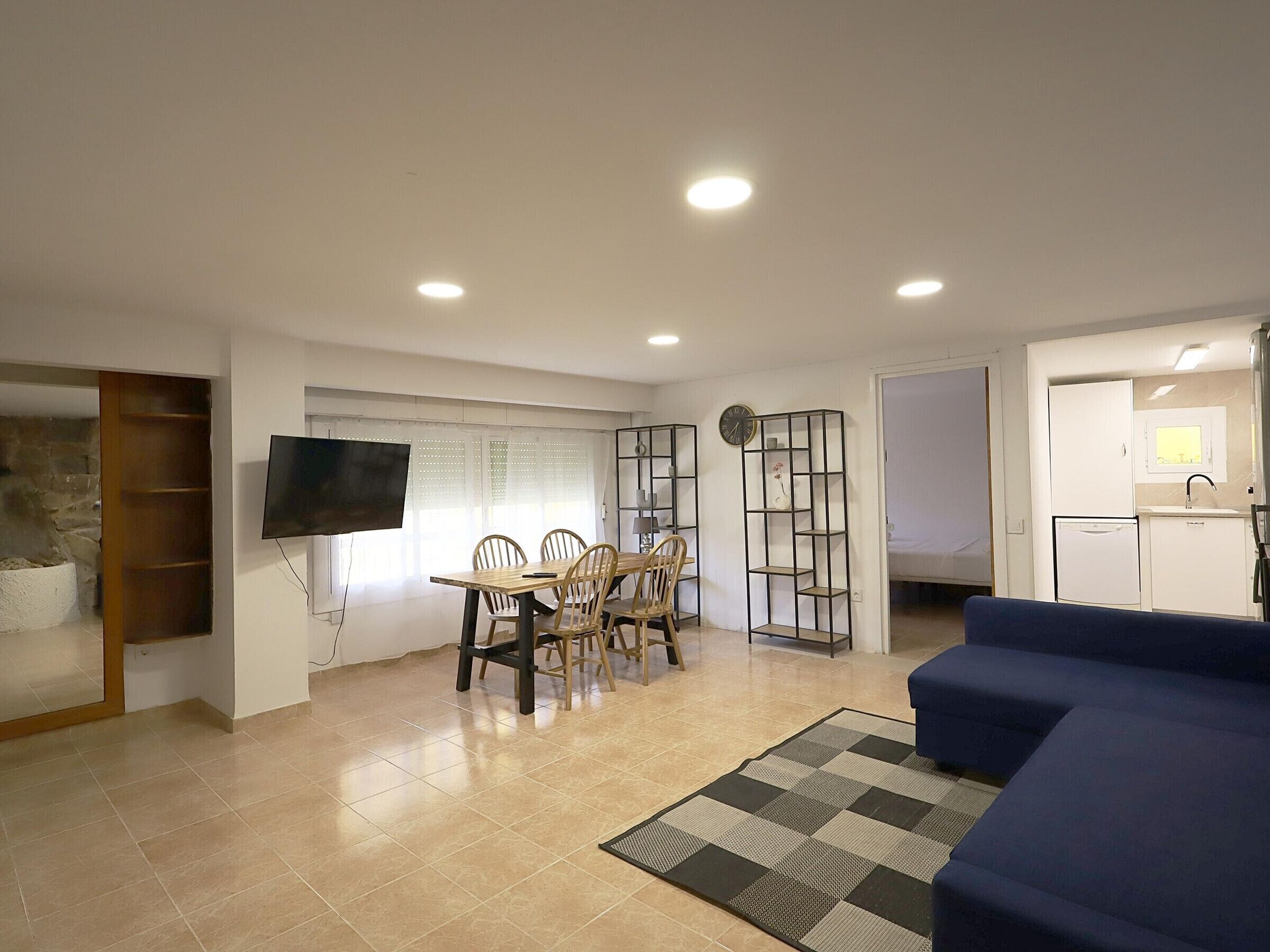 A living room with a dining table, chairs, a TV, a sofa, and a kitchen area with a refrigerator and sink.
