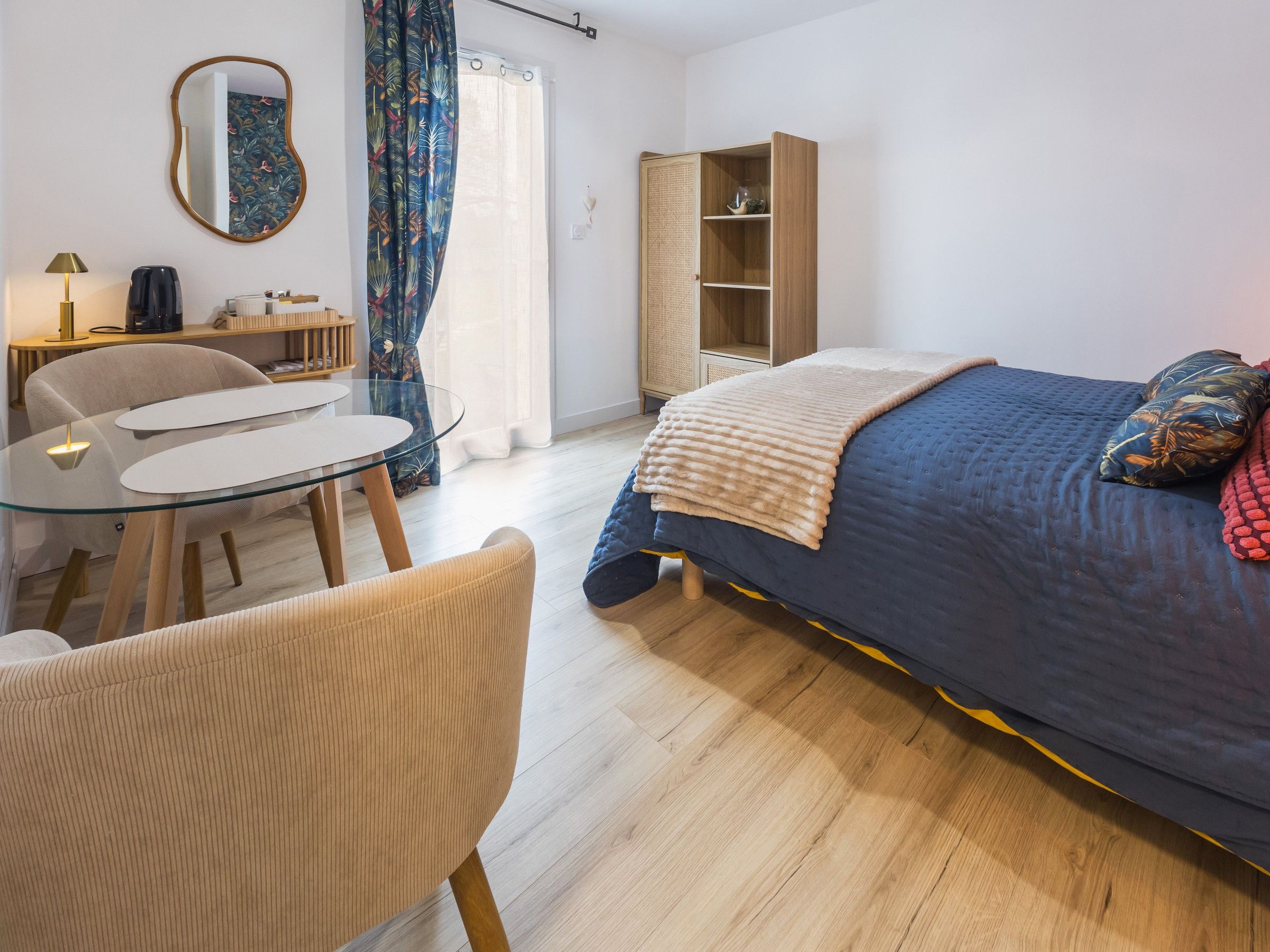 Une chambre à coucher comprenant un lit, une table de chevet, une chaise, une petite table, un miroir, une armoire et une fenêtre avec des rideaux.