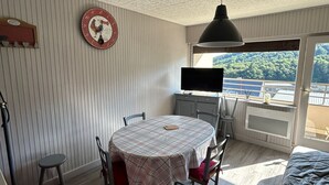 Ein Zimmer mit einem runden Tisch, Stühlen, einem Fernseher, einer Uhr und Blick auf einen Balkon mit Geländer.
