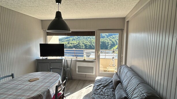 Ein Zimmer mit Esstisch, einer Couch, einem Fernseher und einem Balkon mit Blick auf Grünflächen.