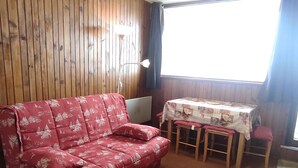Ein Zimmer mit einer holzverkleideten Wand, einem roten Sofa mit floralem Bezug, einem kleinen Tisch mit gemustertem Tischläufer und einem Fenster mit Vorhängen.