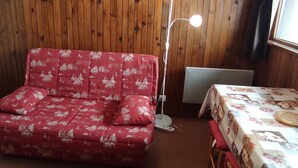 Ein Zimmer mit einem roten, gemusterten Sofa, einer stehenden Stehlampe, einem Tisch mit einer gemusterten Tischdecke und einem Heizkörper.