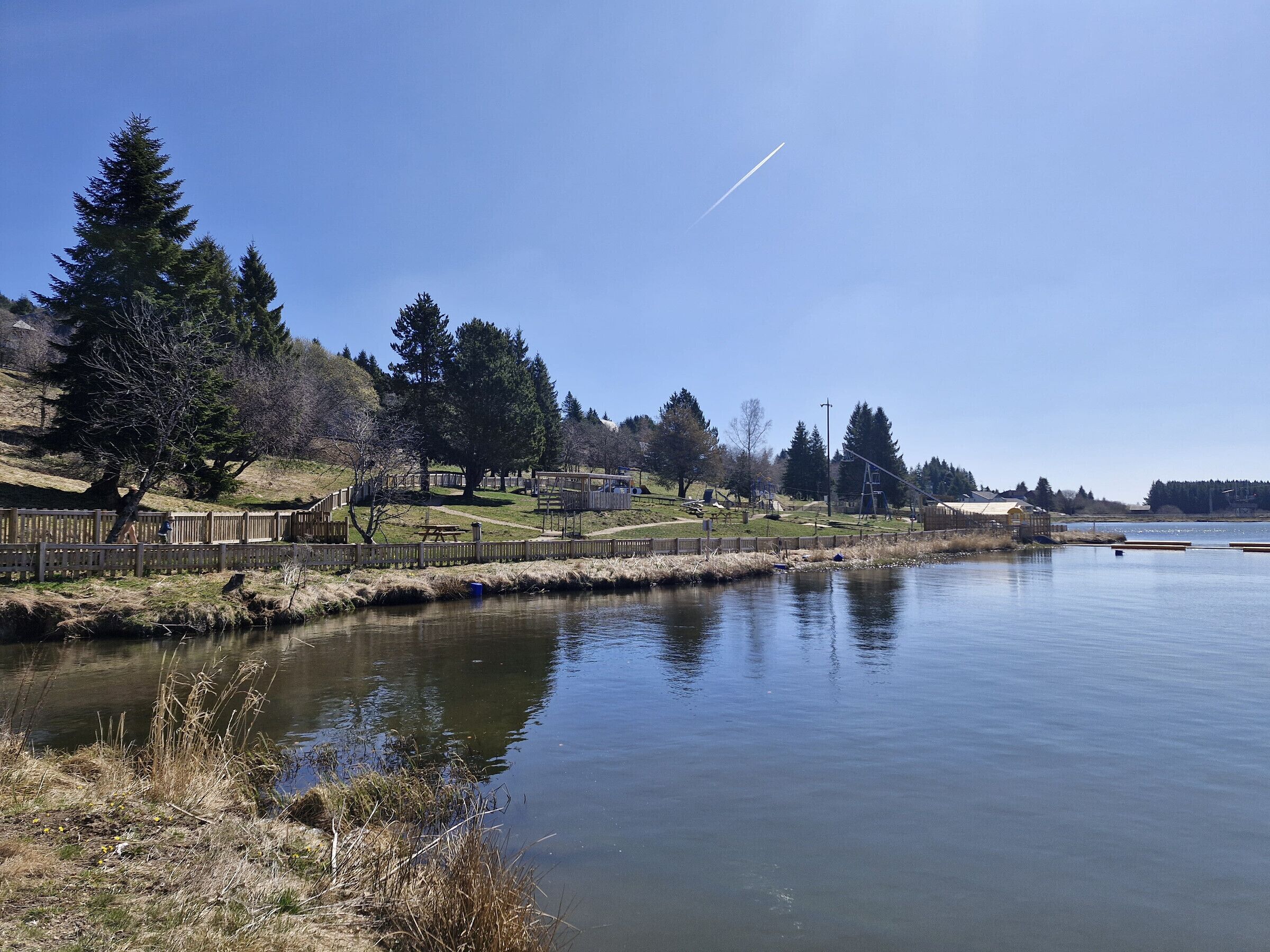A calm lake with a wooden fence and picnic tables along the shore, surrounded by trees and a clear sky.