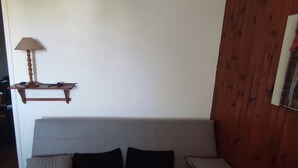 Un sofá gris con dos cojines negros y dos blancos, un estante de madera con una lámpara, y un panel de pared de madera.