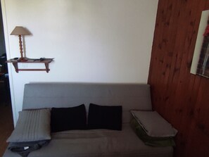 A grey sofa with two black and two white cushions, a small wooden shelf with a lamp, and a wooden wall panel.