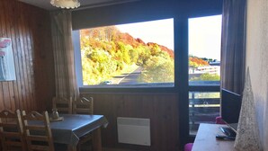 Una habitación con paredes de madera, una mesa con sillas, un radiador y una ventana que da a árboles y una carretera.