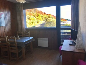 Quarto com decoração em madeira, uma mesa com cadeiras, um radiador e uma janela com vista para árvores e uma estrada.