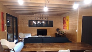 A modern living room with a wooden ceiling, a large wooden dining table, a TV, and a sofa.