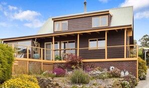 Une maison à deux étages dotée d’un balcon, d’une cheminée et d’un jardin où poussent différentes plantes et fleurs.