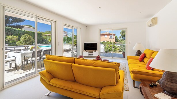 A bright living room with yellow sofas, a TV, and a dining area with a pool view.