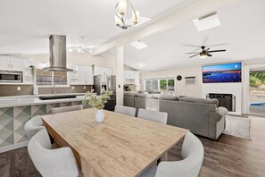 Eine moderne Küche mit weißen Schränken, eine Essecke mit Holztisch und -stühlen und ein graues Sofa in einem Wohnzimmer mit Blick ins Freie.