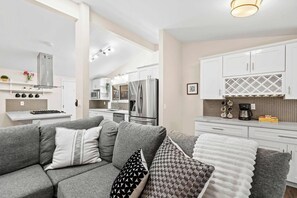 Une cuisine moderne avec des meubles blancs, des appareils en acier inoxydable et un canapé gris agrémenté de coussins à motifs.