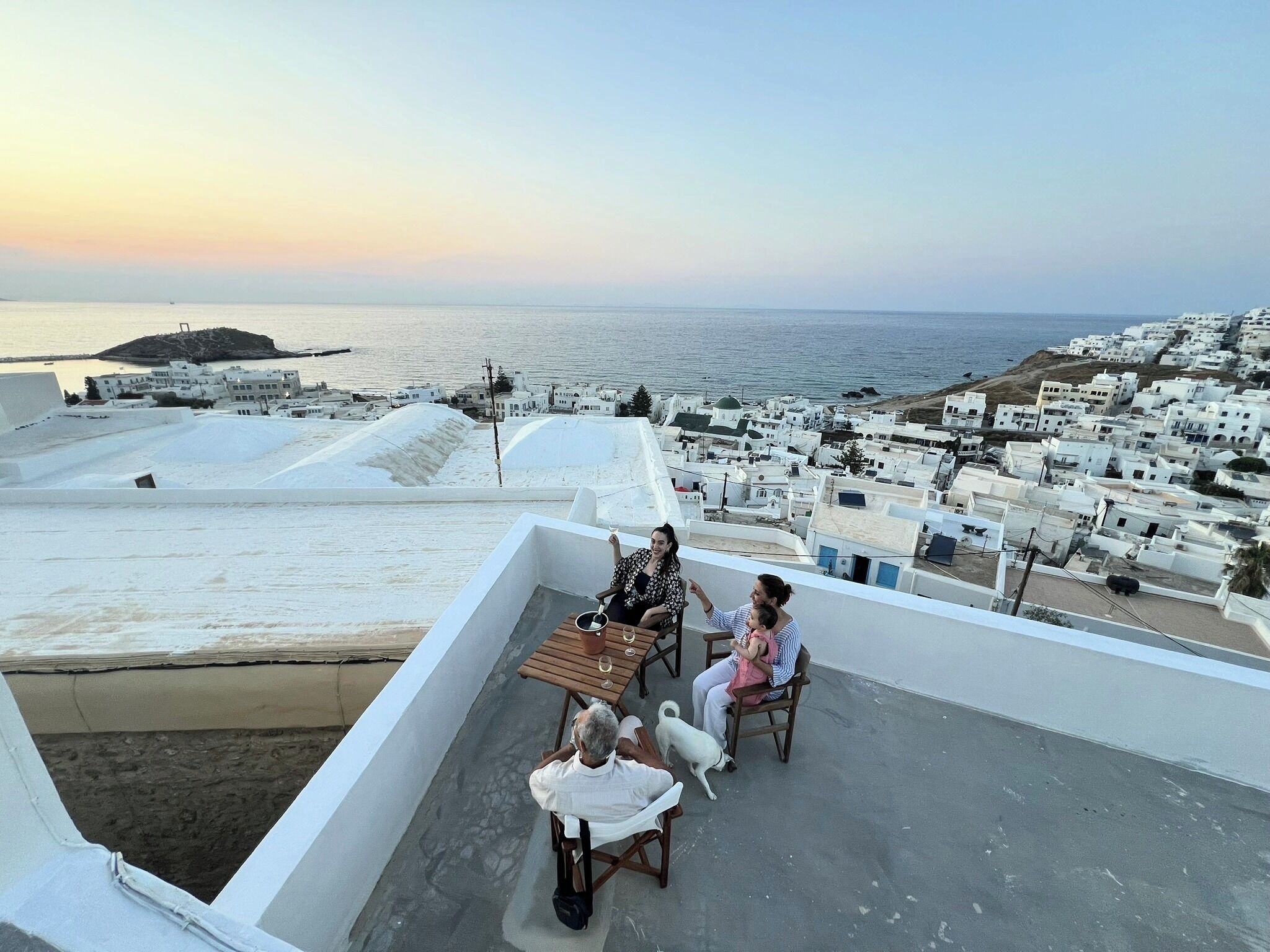 A terrace with a view of a coastal town, a table with chairs, and a white building in the background.
