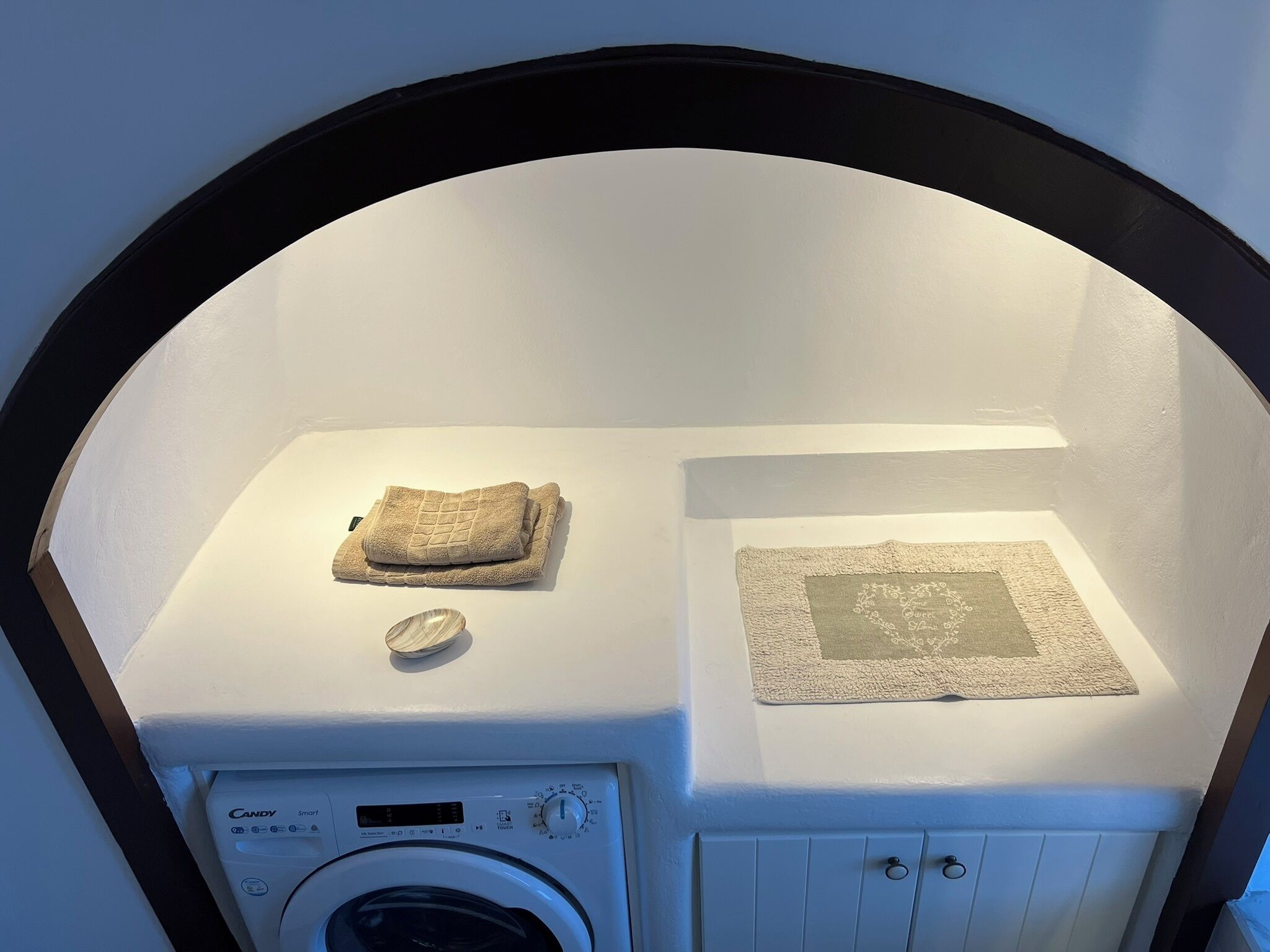 Un espacio de lavandería compacto con una lavadora Candy Smart, una toalla doblada y un felpudo decorativo.