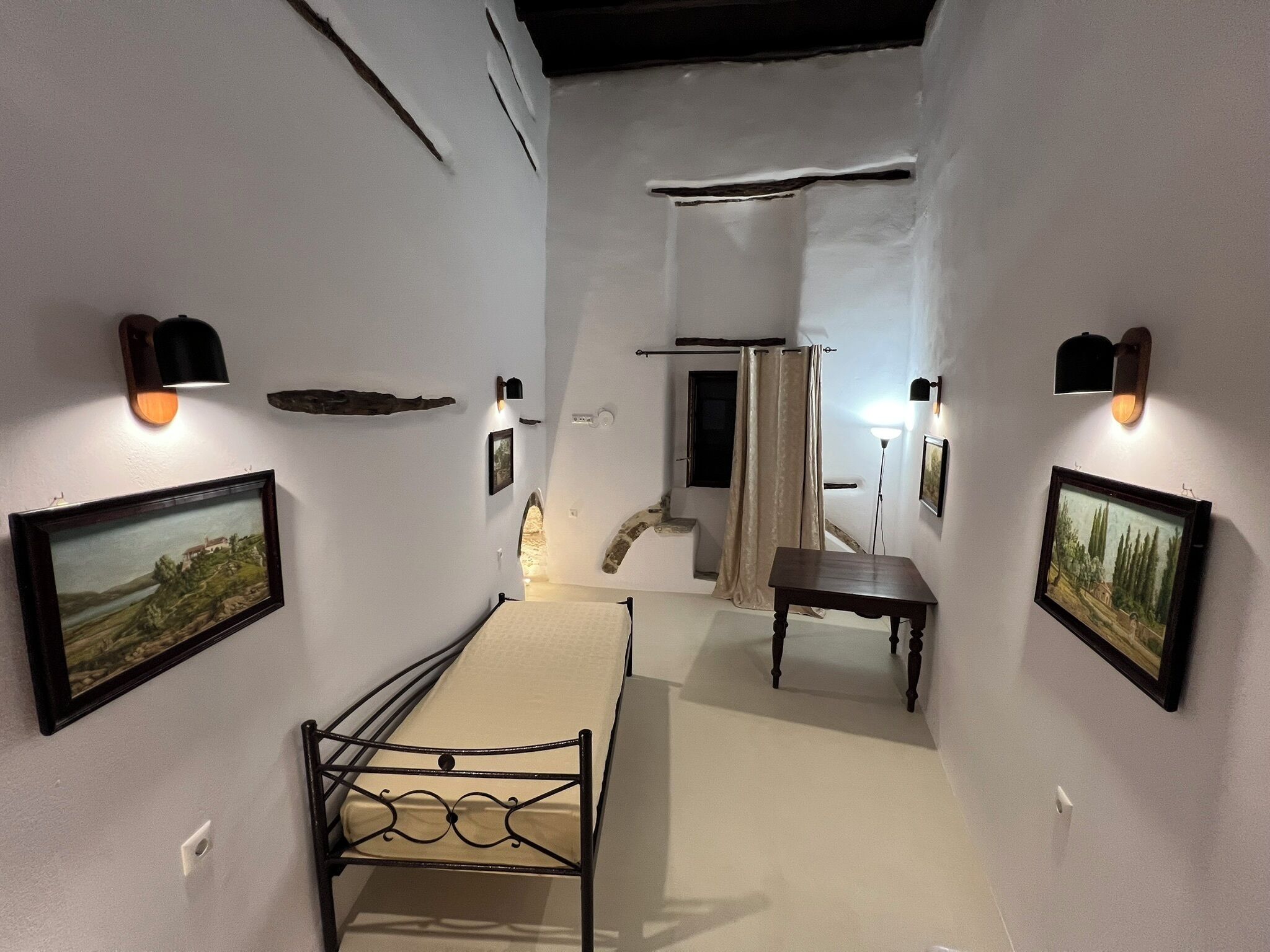 Ein schmaler Flur mit einem Bett, einem kleinen Tisch und zwei Wandleuchten. An den Wänden hängen Bilder in Rahmen und ein Fenster mit Vorhang.