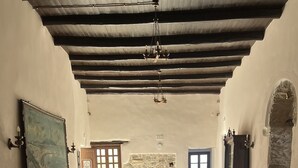 Una habitación amplia con techo alto, una mesa de madera y bancos. Hay una pared de piedra y una puerta que conduce a otra habitación.