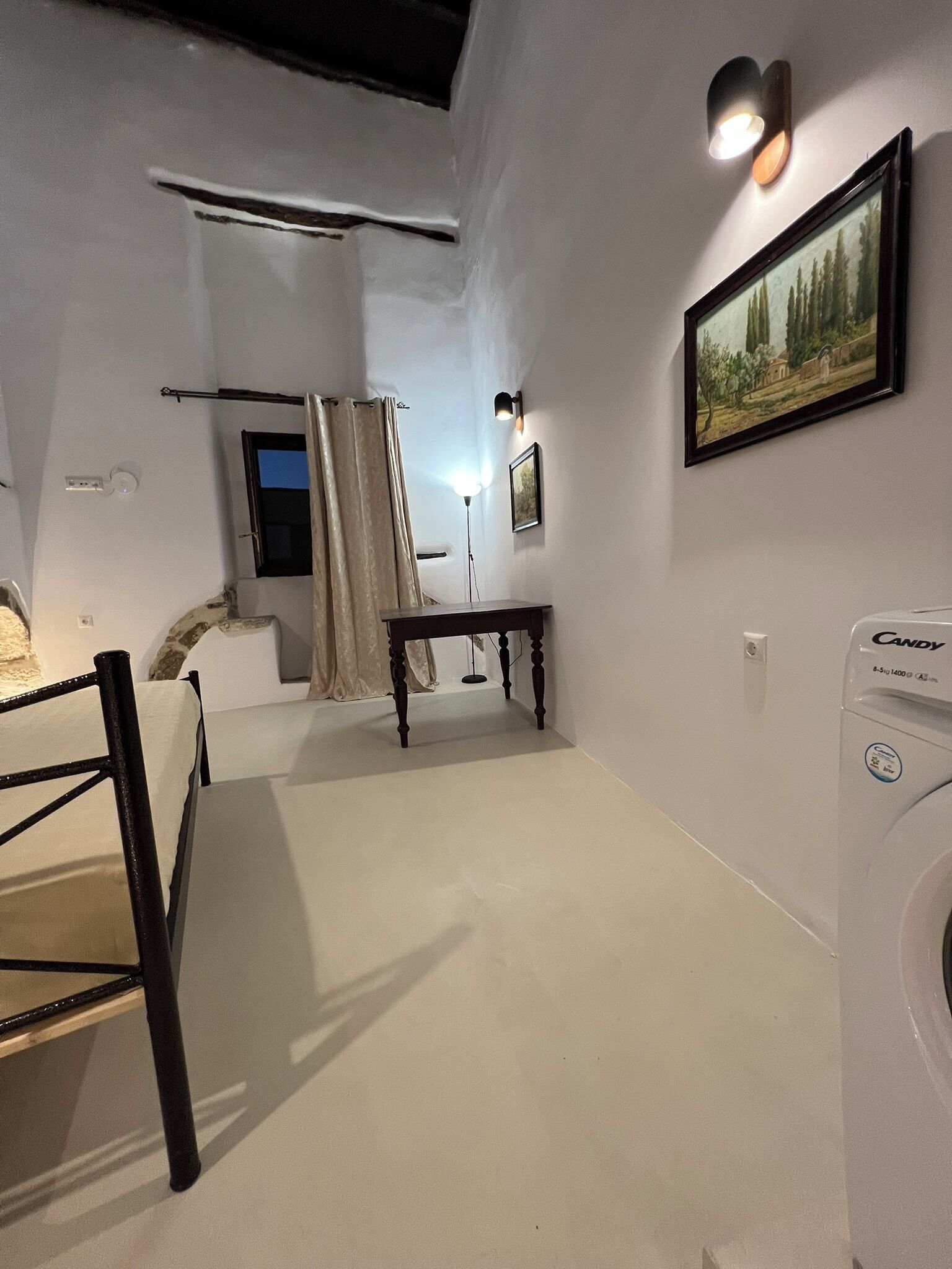 A bedroom with a bed, a table, a chair, a washing machine, a painting, and a window with curtains.
