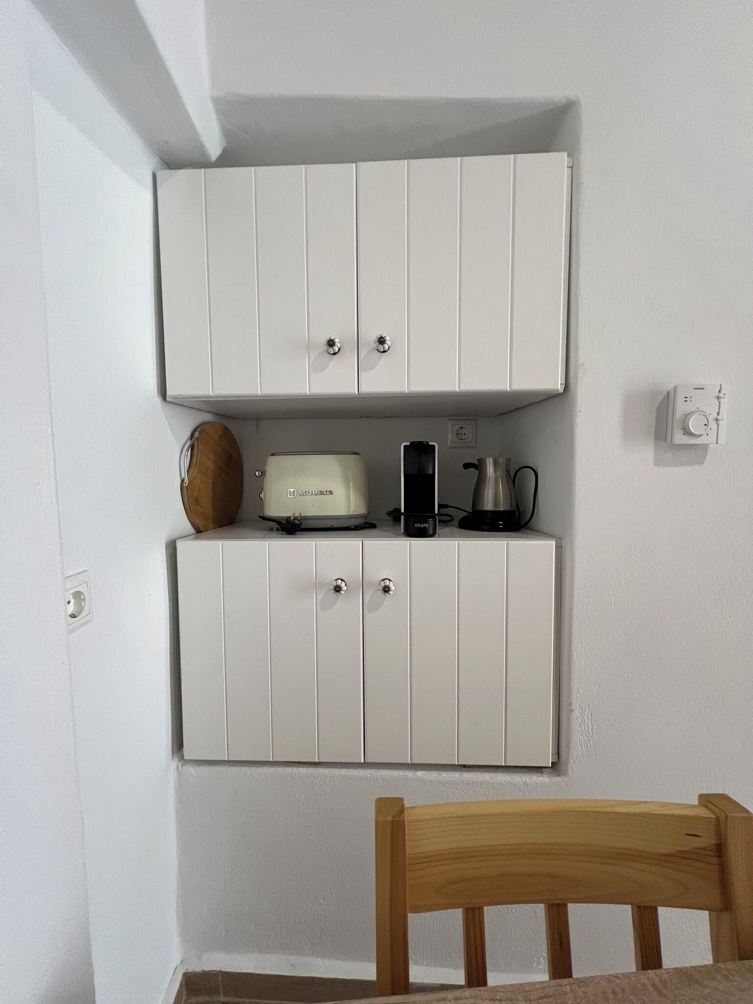 Un mobile bianco con due ante, una macchina da caffè, un bollitore e un telefono sopra.