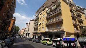 A multi-story yellow building with balconies, parked cars, and trash bins on the street. - ROOMS MONTEVERDE 2 (Rome)
