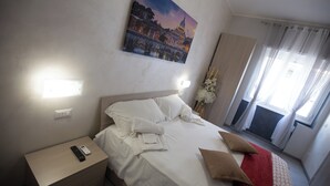 A hotel room with a bed, a nightstand with a remote control, a painting of a cityscape, and a window with curtains. - ROOMS MONTEVERDE 2 (Rome)