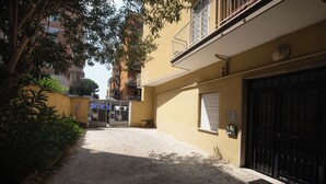 A narrow alleyway with a yellow building, a black gate, and a tree providing shade. - ROOMS MONTEVERDE 2 (Rome)