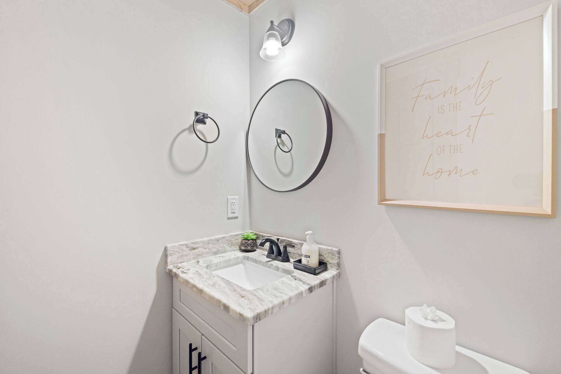 A bathroom with a marble sink, two wall-mounted rings, a toilet, and a framed quote on the wall.