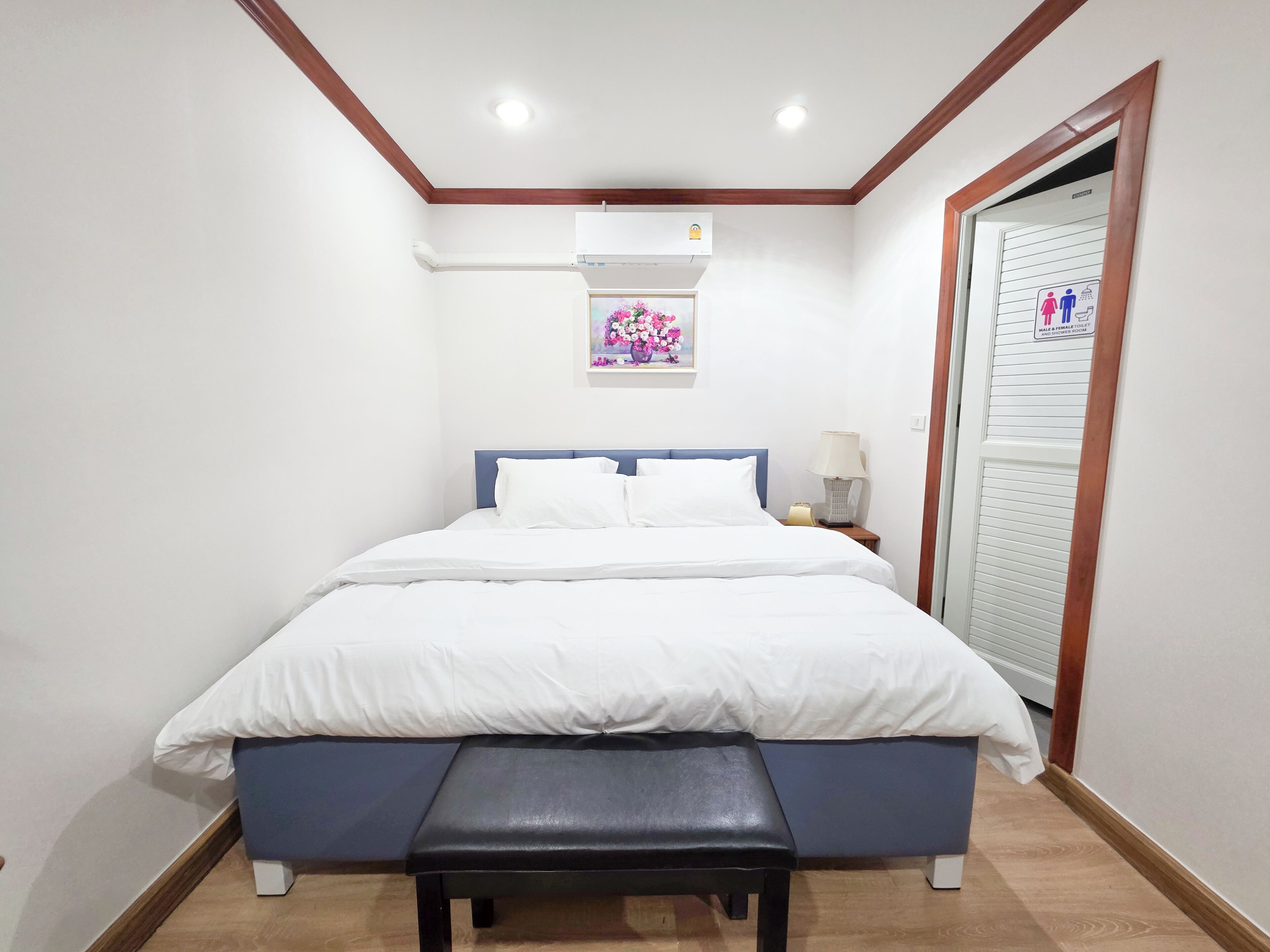 A bedroom with a bed, a bench, an air conditioning unit, a nightstand, and a door with a sign.