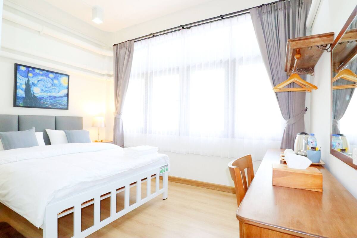 A hotel room with a bed, a desk, a chair, a painting on the wall, and a large window with curtains.