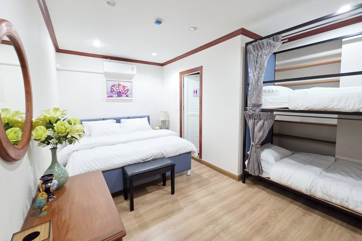 A hotel room with a bed, a bench, a bedside table with a vase, a mirror, a bunk bed, and a wall-mounted air conditioner.