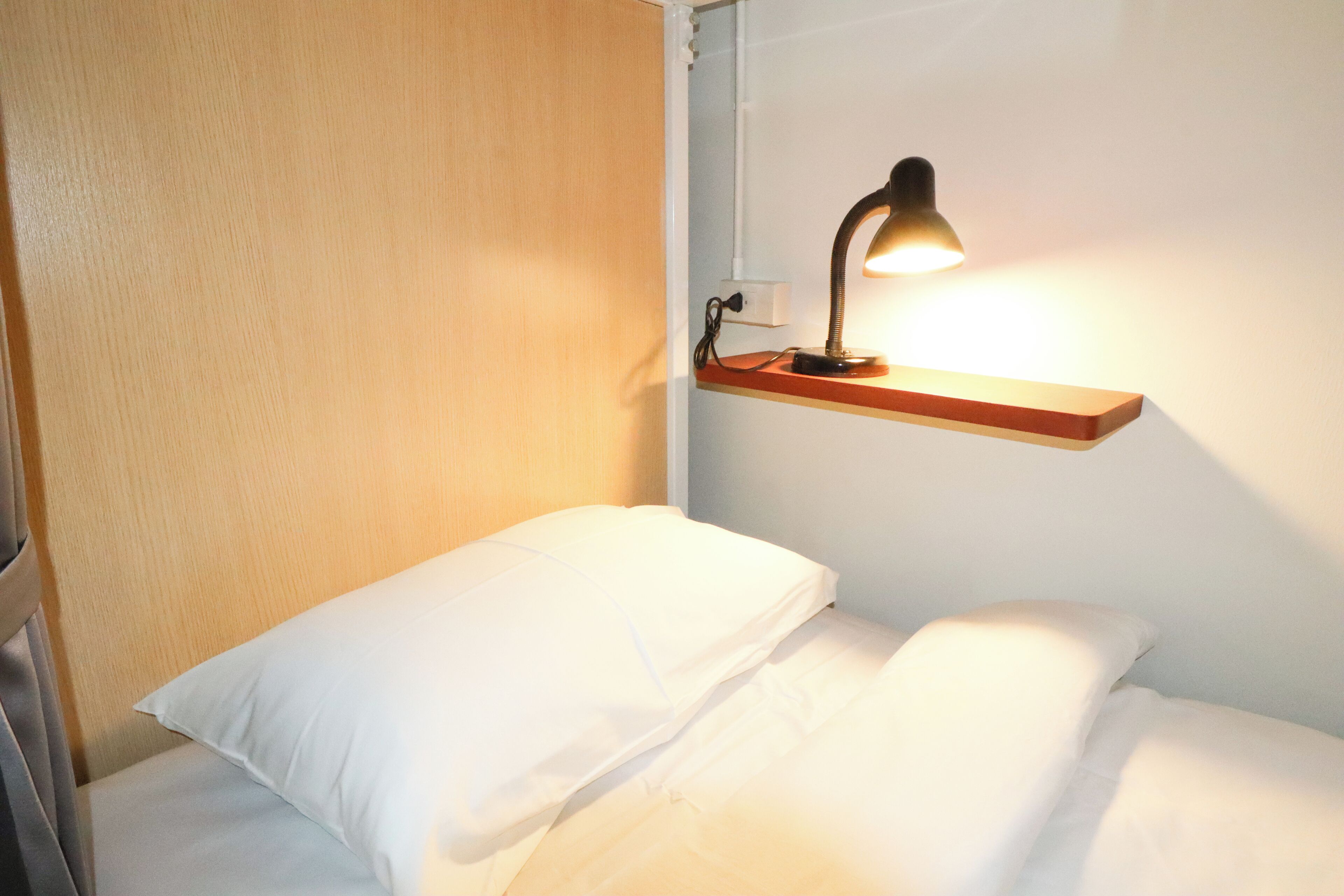 En enkeltseng med hvide sengetøj, et natbord med lampe og en træhylde over sengen.