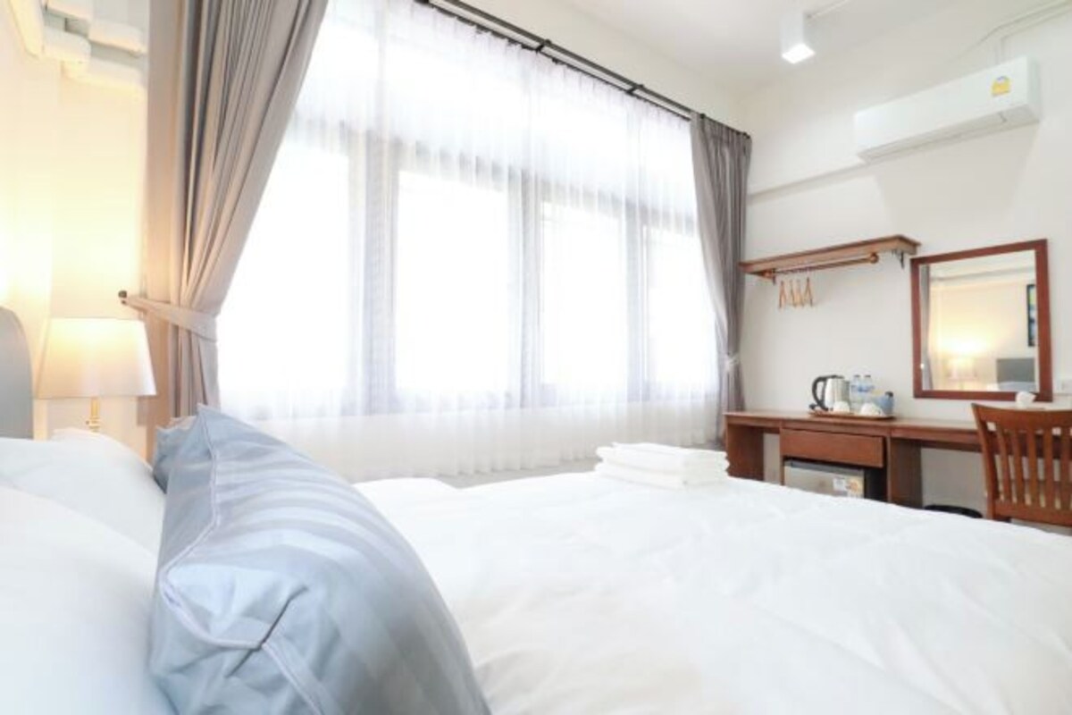 A hotel room with a bed, a desk, a chair, a lamp, a window with curtains, and a mirror.