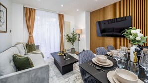A modern living room with a sofa, a coffee table, and a dining area set with plates, glasses, and a vase of flowers. - Modern 4 Bed Flat | Prime Central London Location (Greater London)
