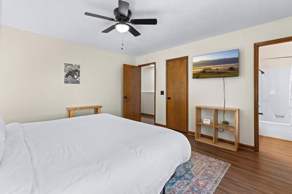 Une chambre avec un grand lit, un ventilateur de plafond, un téléviseur fixé au mur, une étagère en bois et une porte donnant sur une salle de bain.