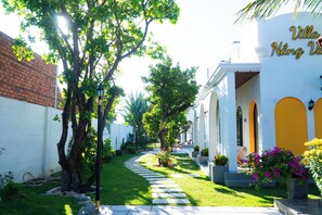 A pathway leading to a building with a white exterior and yellow accents, surrounded by lush greenery and trees. - Green Space Homestay (Ham Thang)