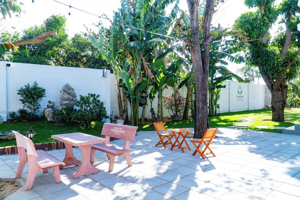 An outdoor patio with a table and chairs, surrounded by lush greenery and a white wall. - Green Space Homestay (Ham Thang)
