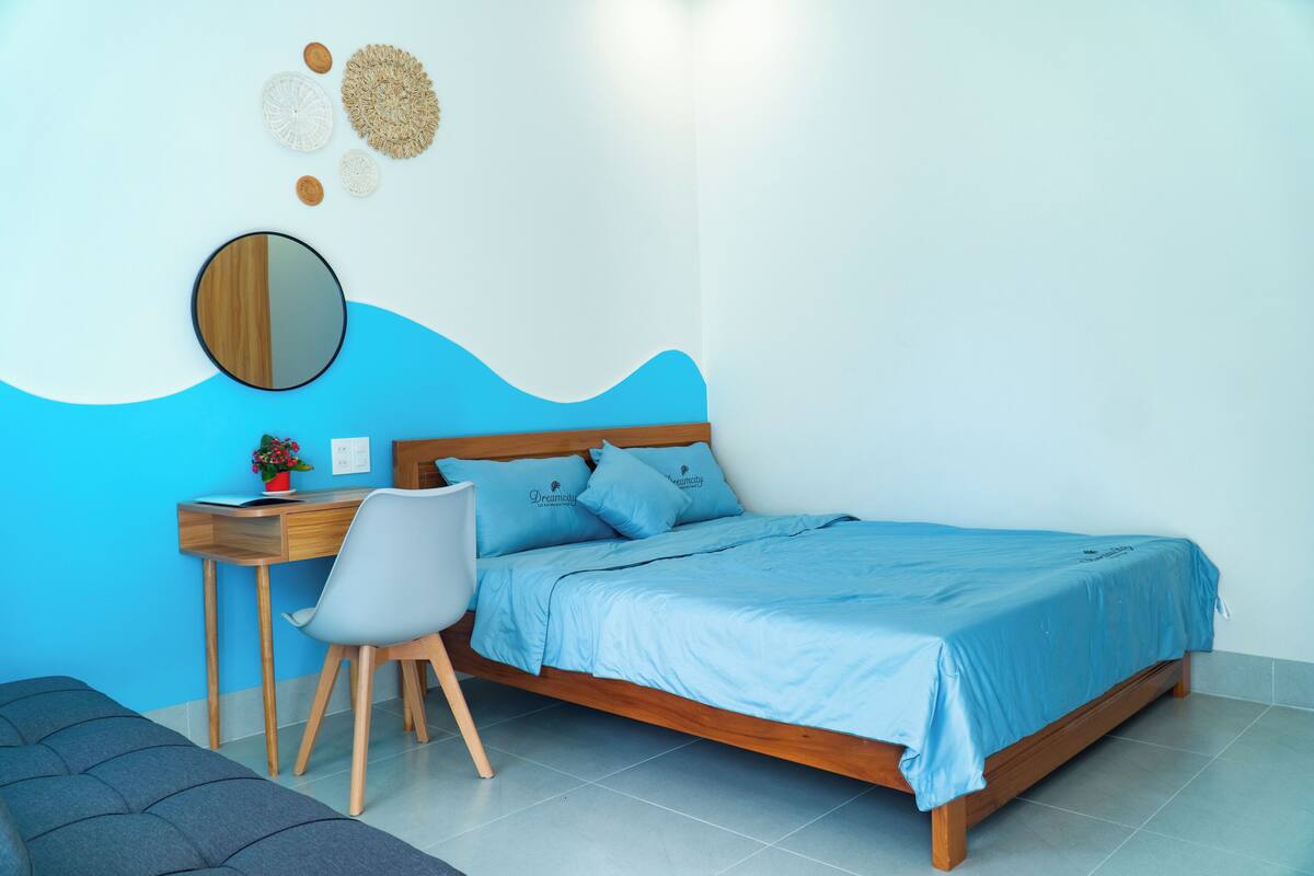 A bedroom with a wooden bed, a desk with a chair, a small table with a plant, and a mirror on the wall.