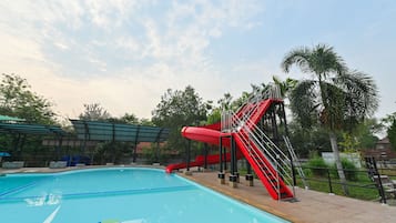 A swimming pool with a red slide and stairs, surrounded by palm trees and a covered area.