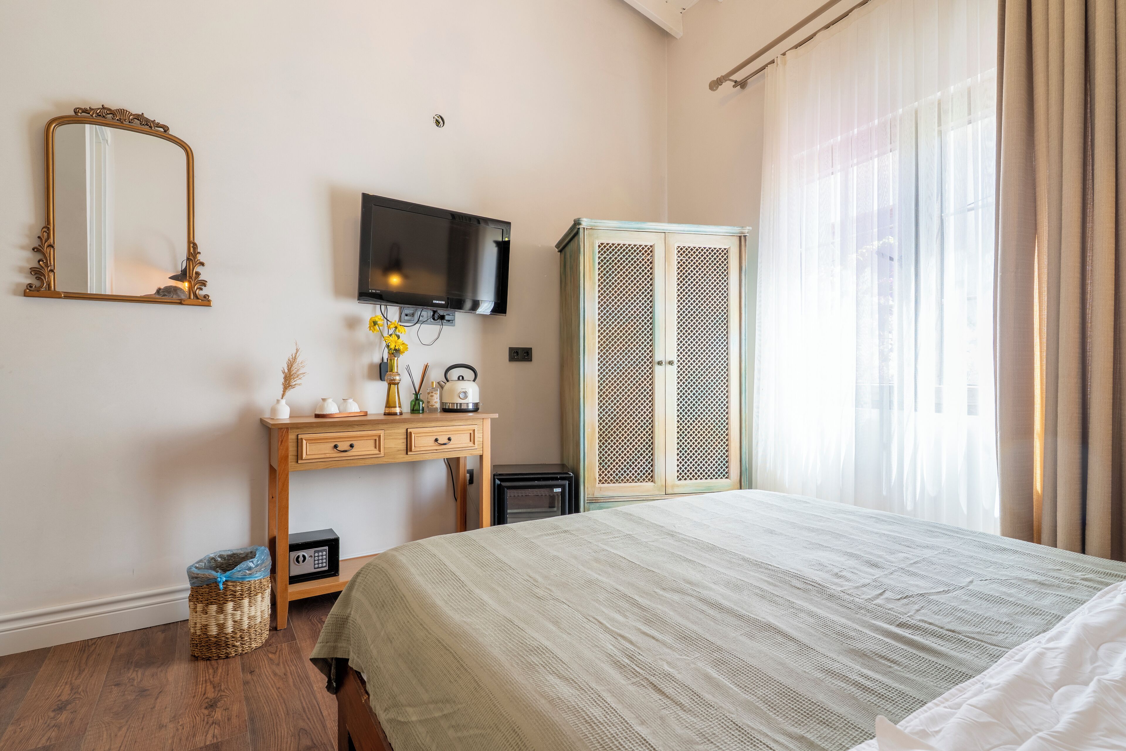 A bedroom with a bed, a TV mounted on the wall, a wardrobe, a desk with a mirror, and a window with curtains.