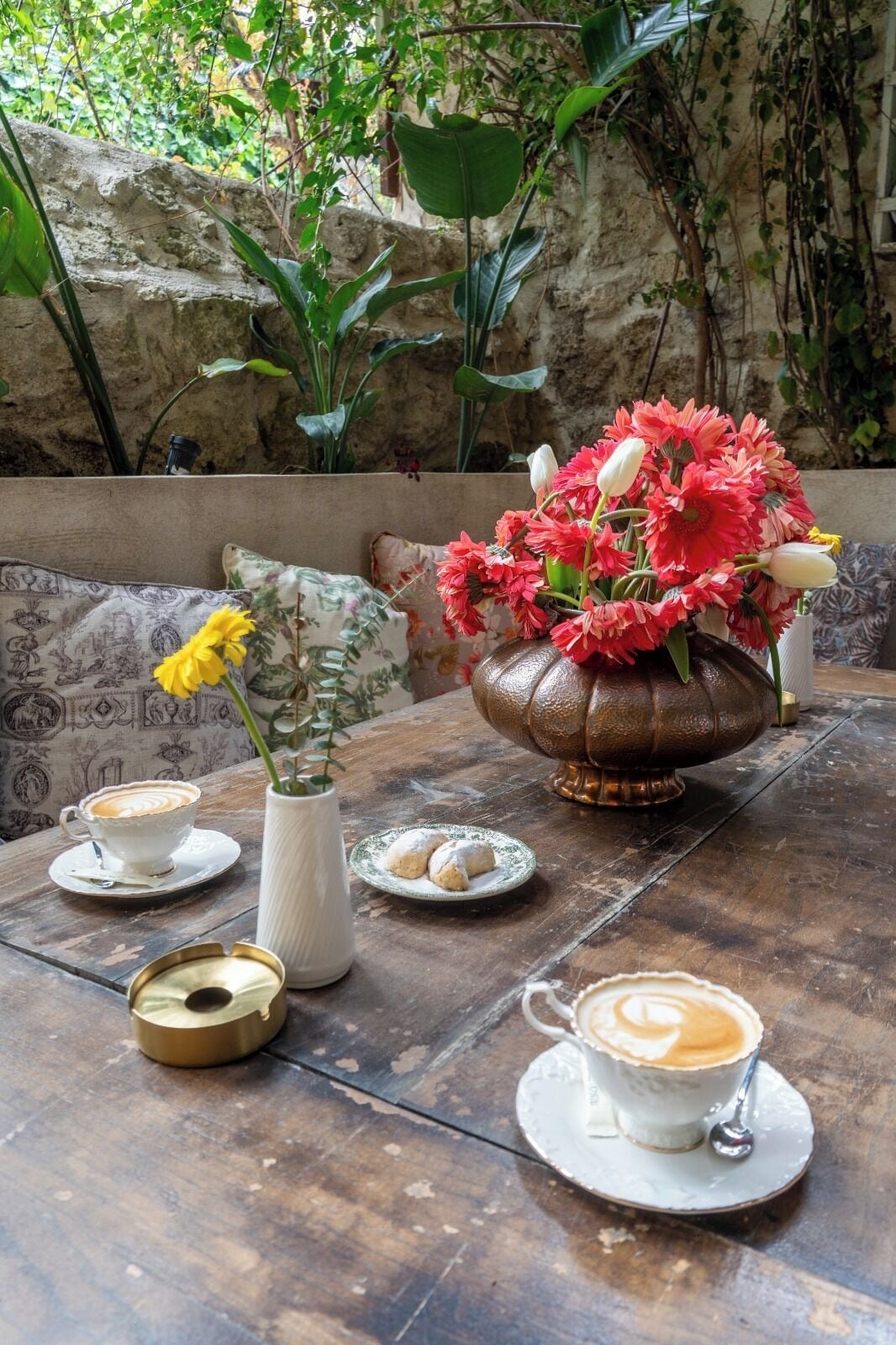 A rustic outdoor seating area with a table set for coffee and pastries, surrounded by greenery and a stone wall.